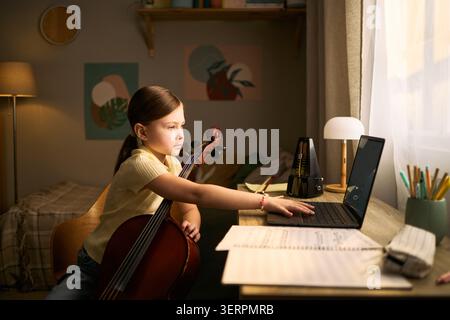 Child girl studying music and practicing cello while using laptop at desk, supporting online music lessons and home education marketing for learning platforms Stock Photo