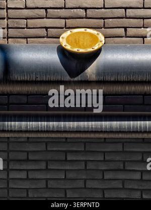 Close-up of horizontal steel pipeline affixed to textured brick wall featuring circular brass with bolts, sunlight casting sharp shadow across pipe Stock Photo