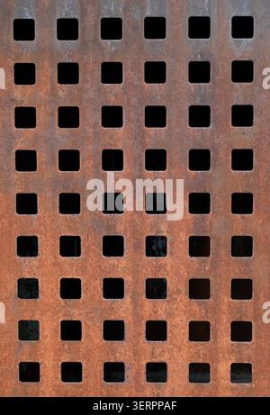 Vertical close up of a weathered rusty metal surface with a grid of square cut outs, dark openings, pitted corrosion, oxidized texture Stock Photo