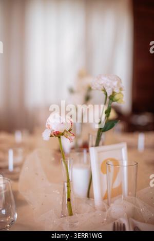 Delicate close-up of an elegant wedding table setting featuring a fresh white-pink peony in a slim glass vase, soft sparkling tulle, crystal glassware Stock Photo