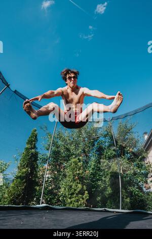 Young man jumping high on a backyard trampoline in red shorts under a clear blue sky Stock Photo