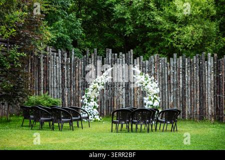 An outdoor wedding ceremony setup features dark chairs, a lush white floral arch, and a rustic log fence surrounded by green trees Stock Photo
