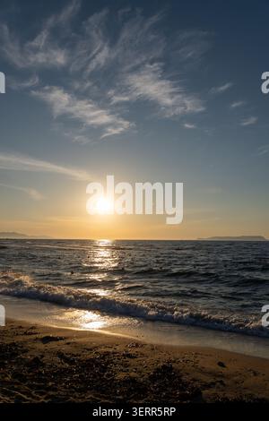A vertical orientation photograph showing a peaceful golden sunset over the ocean. Beautiful wispy clouds spread across the blue sky while the sun ref Stock Photo
