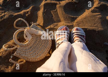 A first-person perspective view of a woman standing on the sandy beach next to a stylish knitted straw bag with a lace ribbon. The low golden sunset l Stock Photo