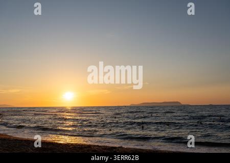 A panoramic seascape featuring a bright golden sun setting over the calm ocean horizon. A brilliant path of warm light reflects on the rippling water Stock Photo