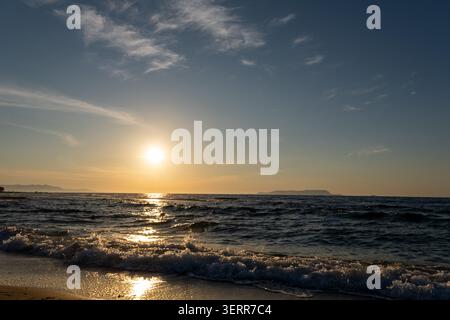 A beautiful sunset over the sea with a bright golden sun path reflecting on the water surface. Gentle ocean waves with white foam crash on the sandy b Stock Photo