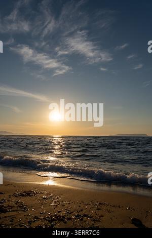 A vertical orientation image capturing the golden sun low over the sea, casting bright reflections on the water. Gentle sea foam from breaking waves w Stock Photo