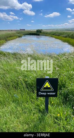 A sign warns of deep water to prevent accidental drowning during hot weather & heatwaves near a small lake at Whitby Abbey in North Yorkshire Stock Photo