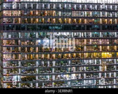 Aerial view of illuminated office windows in a modern glass skyscraper at night showing desks, plants and people working in Poland. Stock Photo