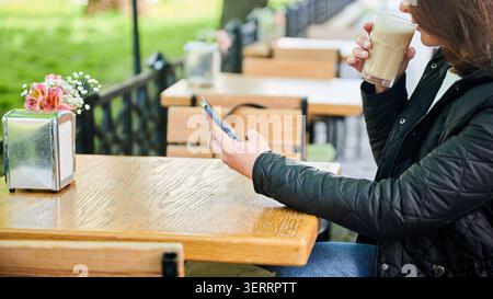 Young woman drinking latte while using smartphone at outdoor cafe, sitting at wooden table, wearing casual jacket, relaxing alone near green park in soft daylight Stock Photo