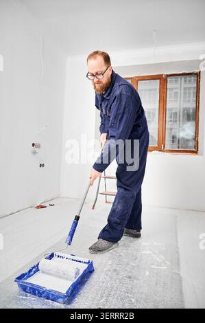 Builder applying paint layer on floor using roller and tray during renovation. Fresh white finish for smooth surface, repair work and residential upgrade concept Stock Photo