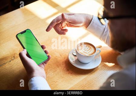 Man using smartphone with green screen while drinking cappuccino in cafe, pointing at mobile display, latte art coffee on table, modern lifestyle, technology and communication concept Stock Photo