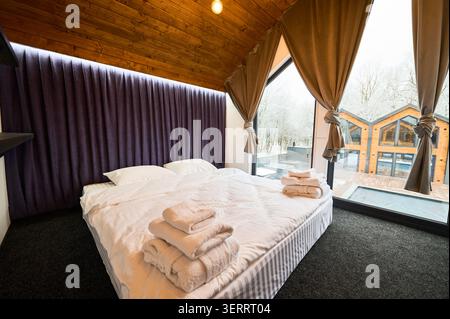 Cozy bedroom with soft bedding and towels. Wooden ceiling and pendant lights, large window facing snowy yard and cabins, peaceful rest and holiday rental concept Stock Photo