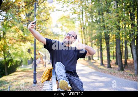 White man taking selfie on bench smiling and giving thumbs up while holding phone on stick, wearing glasses and beard, black shirt, paved park path with lamp posts and golden autumn foliage, candid Stock Photo