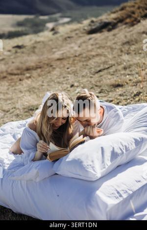 Happy couple reading book together outdoors on white bedding in mountains Stock Photo