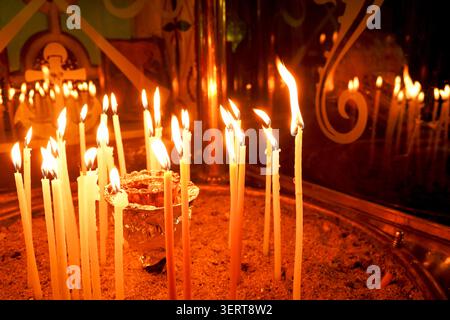 Atmospheric low-light shot of numerous slender golden wax candles burning in a sand-filled container inside a traditional Orthodox church. Stock Photo