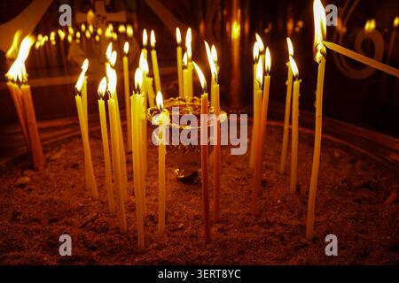 Atmospheric low-light shot of numerous slender golden wax candles burning in a sand-filled container inside a traditional Orthodox church. Stock Photo