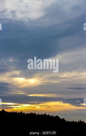 Stunning sunset sky with dramatic blue-grey clouds and golden sunlight piercing through the horizon over a dark, silhouetted forest treeline. Stock Photo