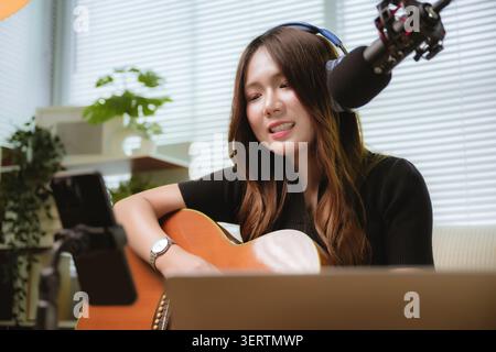 Asian young woman musician singing playing acoustic guitar while live streaming video content, creator home studio with microphone laptop smartphone c Stock Photo