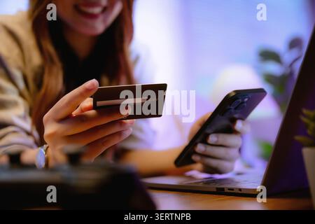 Person holds credit card and smartphone near laptop. smile shows happiness in online shopping activity. Perfect for payment pages or shopping website. Stock Photo