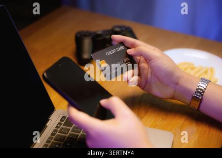 Close-up hands holds credit card and smartphone near laptop and game controller, prepare buying digital game items. Technology online shopping, credit Stock Photo