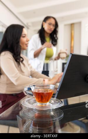 Two women consulting and working at home workspace with glass teacup on saucer and laptop. Collaboration, planning, bright, minimalist, kitchen, offic Stock Photo
