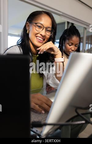 Female coworkers wearing glasses smiling and collaborating at desk in office with laptops, monitor Stock Photo