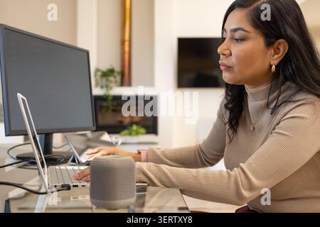 Female in beige turtleneck sitting at glass desk at home workspace, typing on laptop with monitor Stock Photo