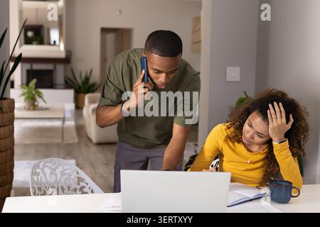 Diverse couple using open laptop at dining table, man leaning talking on phone, woman writing notes Stock Photo