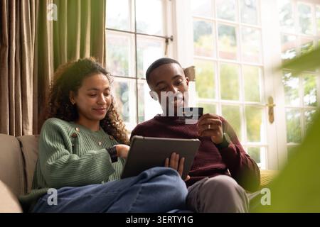 Couple sitting on beige sofa at home sharing tablet, woman in green sweater, man holding card Stock Photo