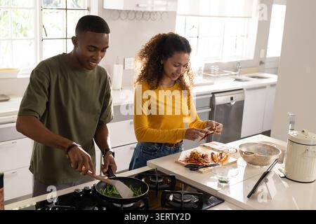 Diverse couple stirring green beans and peeling veggies at central island in modern kitchen, tablet Stock Photo