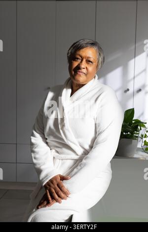 Senior African American woman sitting on vanity in white terry robe, green plant, blind shadows Stock Photo