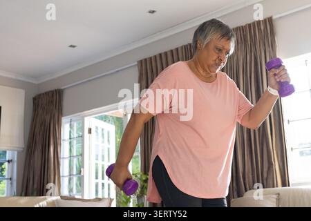 Senior African American woman in pink top exercising with purple weights in living room, copy space Stock Photo