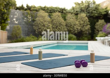 Three dark blue yoga mats are lying on wooden deck by pool, with bottles and dumbbells Stock Photo