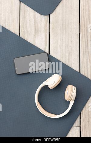 Beige wireless headphones are resting on dark blue yoga mat on wooden deck, black smartphone near Stock Photo
