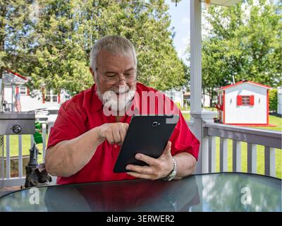 A senior sits at a table on a porch and looks at a tablet. He smiles and points at the screen. The sun is shining, and trees are visible behind him. Small buildings are in the background. Stock Photo