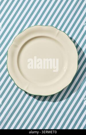 Overhead flat lay shot of an empty minimalist white ceramic plate placed on a rustic,  blue and white striped linen tablecloth. Captured under bright, Stock Photo