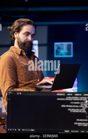 Computer scientist walking in apartment designing and developing software applications using laptop. IT professional standing in living room, implementing and maintaining company systems. Stock Photo