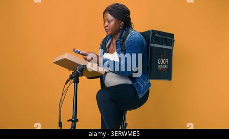 Black woman delivers takeout orders on bicycle, utilizing wireless payment device for seamless transactions. African american delivery person taking pos machine from pocket for contactless pay. Stock Photo