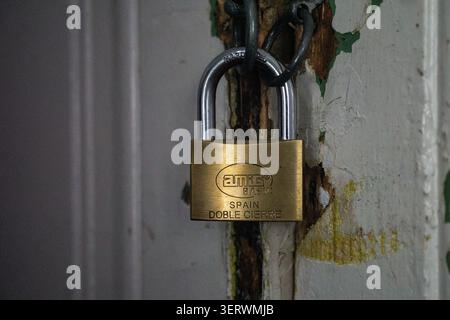 A brass padlock hangs on a weathered door, its surface gleaming against the peeling paint and rusted metal. The lock, marked with "Euthy" and "Spain," Stock Photo