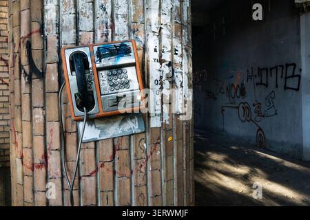 An old payphone mounted on a brick wall covered in graffiti, with a dark alleyway in the background. Stock Photo