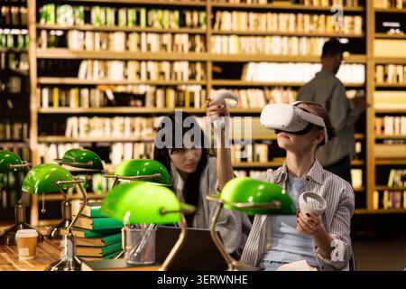 Student using VR device and controller to explore immersive learning at library desk, EdTech and virtual reality used for digital learning and future in education. Modern interactive tools. Stock Photo