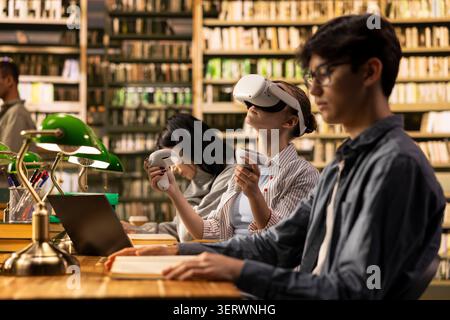 Student wears VR glasses and holds controllers in university library, using immersive technology beside classmates with open books. Virtual reality technology in education, digital learning. Stock Photo