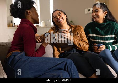 Diverse female friends sitting closely on gray sofa at home holding ceramic mugs, laughing Stock Photo