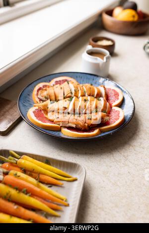 Blue ceramic plate is holding glazed protein slices over grapefruit on countertop with carrots Stock Photo