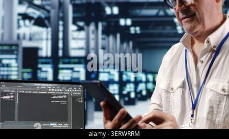 Senior technician in data center, solving tasks on tablet while running scripts on laptop terminal, with skilled technician amidst servers, accessing databases and manipulating lines of code. Stock Photo