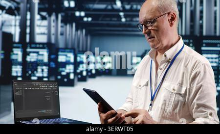 IT solutions expert in data center environment, solving tasks on tablet while running scripts on laptop terminal, with skilled specialist amidst servers, accessing databases. Stock Photo