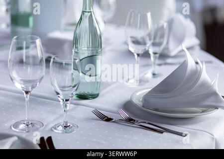 The table is arranged for a meal with glasses and a bottle. White napkins are neatly folded on plates. The setting is inside a restaurant during the e Stock Photo