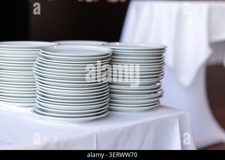 A stack of white plates sits on a table covered with a white cloth in a restaurant. The setting is well organized for a dining event. Natural light fi Stock Photo