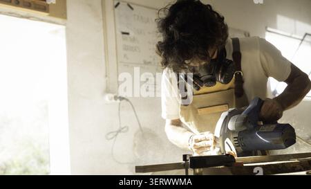 View of a woodworker wearing a protective mask using a circular saw to cut a wooden plank in a workshop in Lisbon, Lisbon, Portugal. Stock Photo